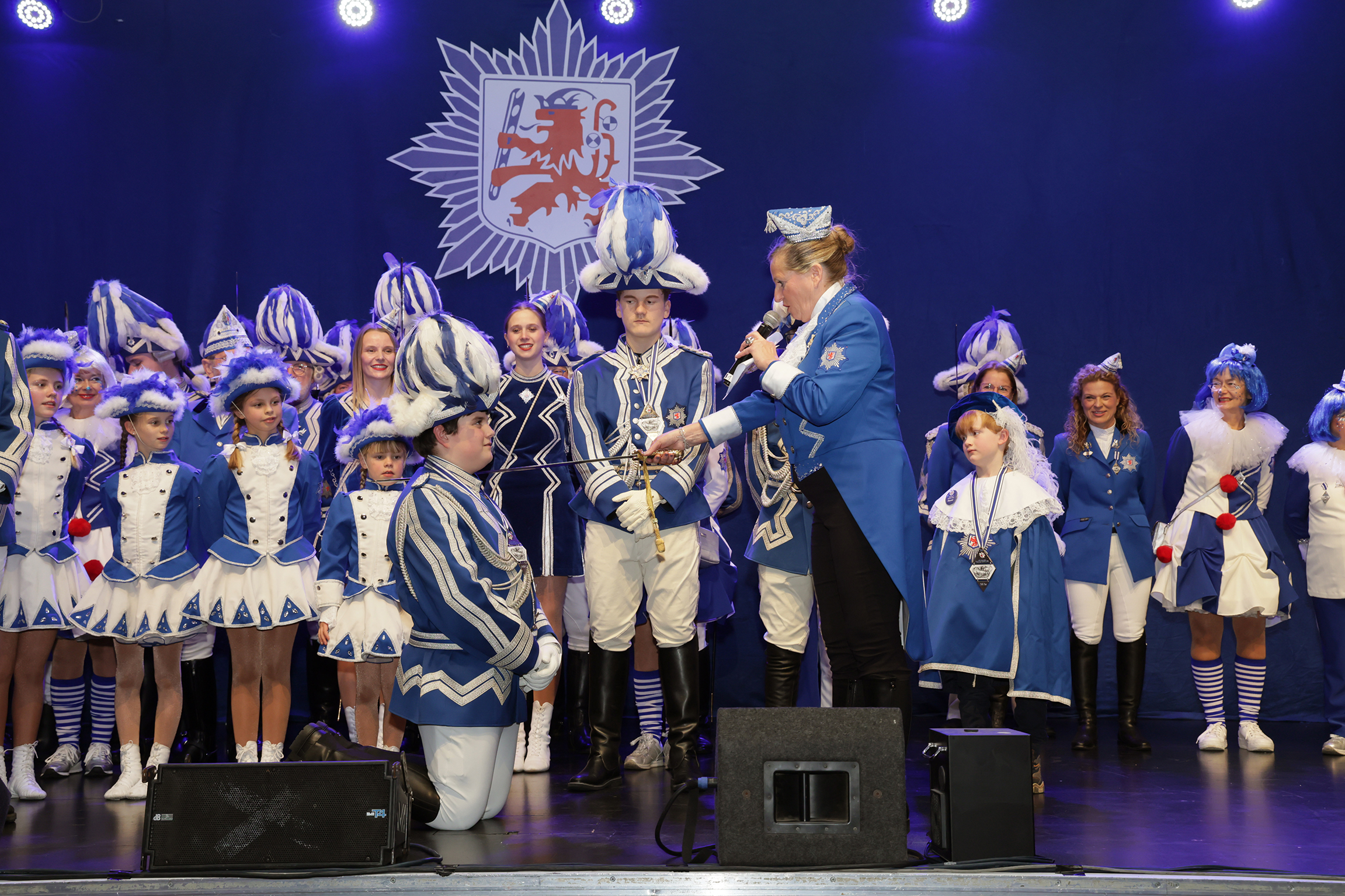 Präsidentin Ute Heierz-Krings in blauem Frack schlägt mit einem Degen den vor ihr knienden Tom Marx in Gardeuniform, umgeben von Kindern und Jugendlichen der Prinzengarde in blau-weissen Uniformen.