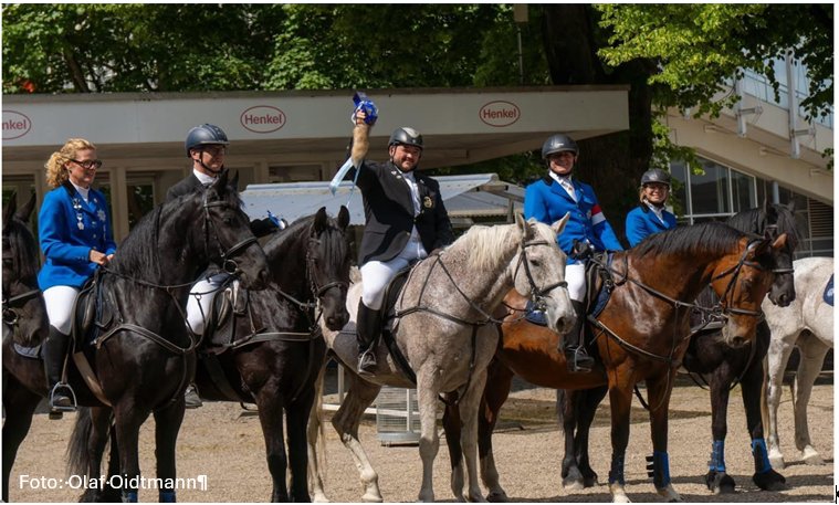 F&uuml;nf Reiter in Turnierkleidung posieren mit ihren Pferden; der Mann in der Mitte h&auml;lt triumphierend eine blaue Schleife hoch.